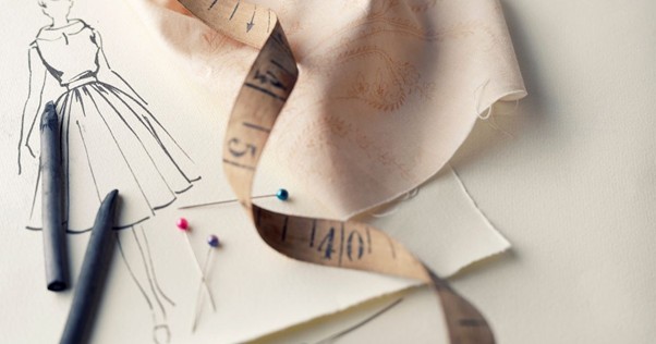 Fashion design workspace featuring a dress sketch, fabric swatch, measuring tape, colored pins, and drawing tools arranged on a light surface.